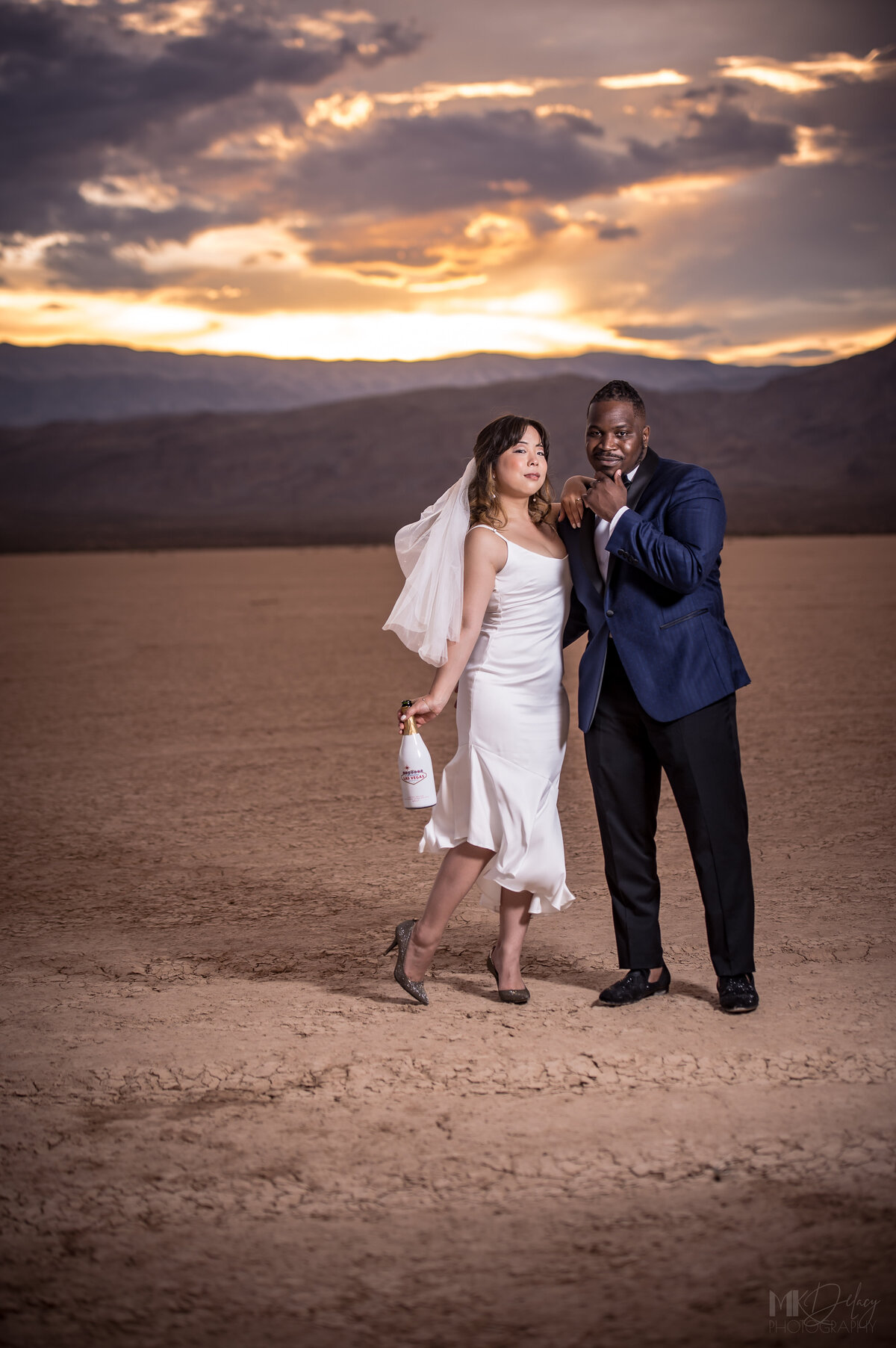 Bride and groom strike a pose in theri dry lake bed elopement portraits as bride holds iconic welcome to las vegas champagne bottle the sun sets over the top of the mountains behind them turning the mountains purple and the clouds from rain las vegas elopement on the dry lake bed  at golden hour groom in blue suit jacket and black  pants  las vegas elopement eloping in vegas  las vegas wedding photographers las vegas wedding photography mk delacy photography