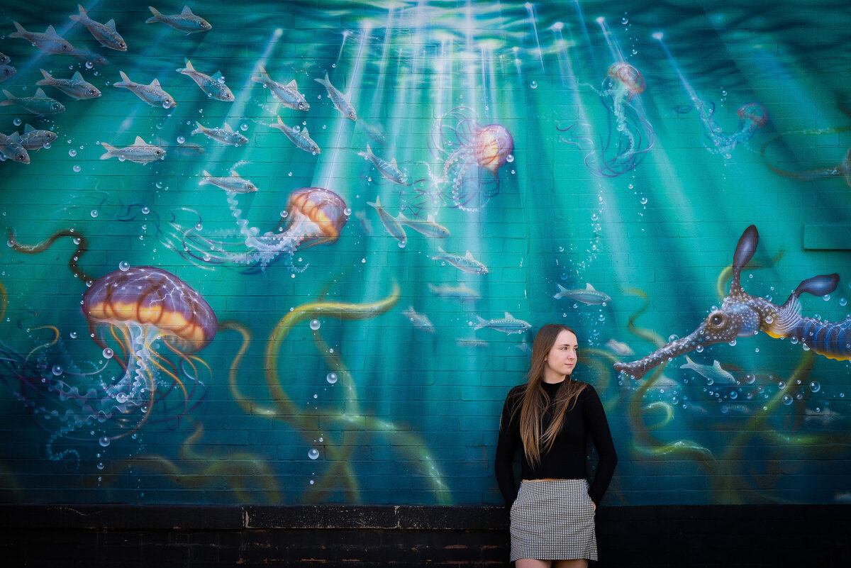 Teenage girl poses for senior portrait in front of wall painted with a seascape
