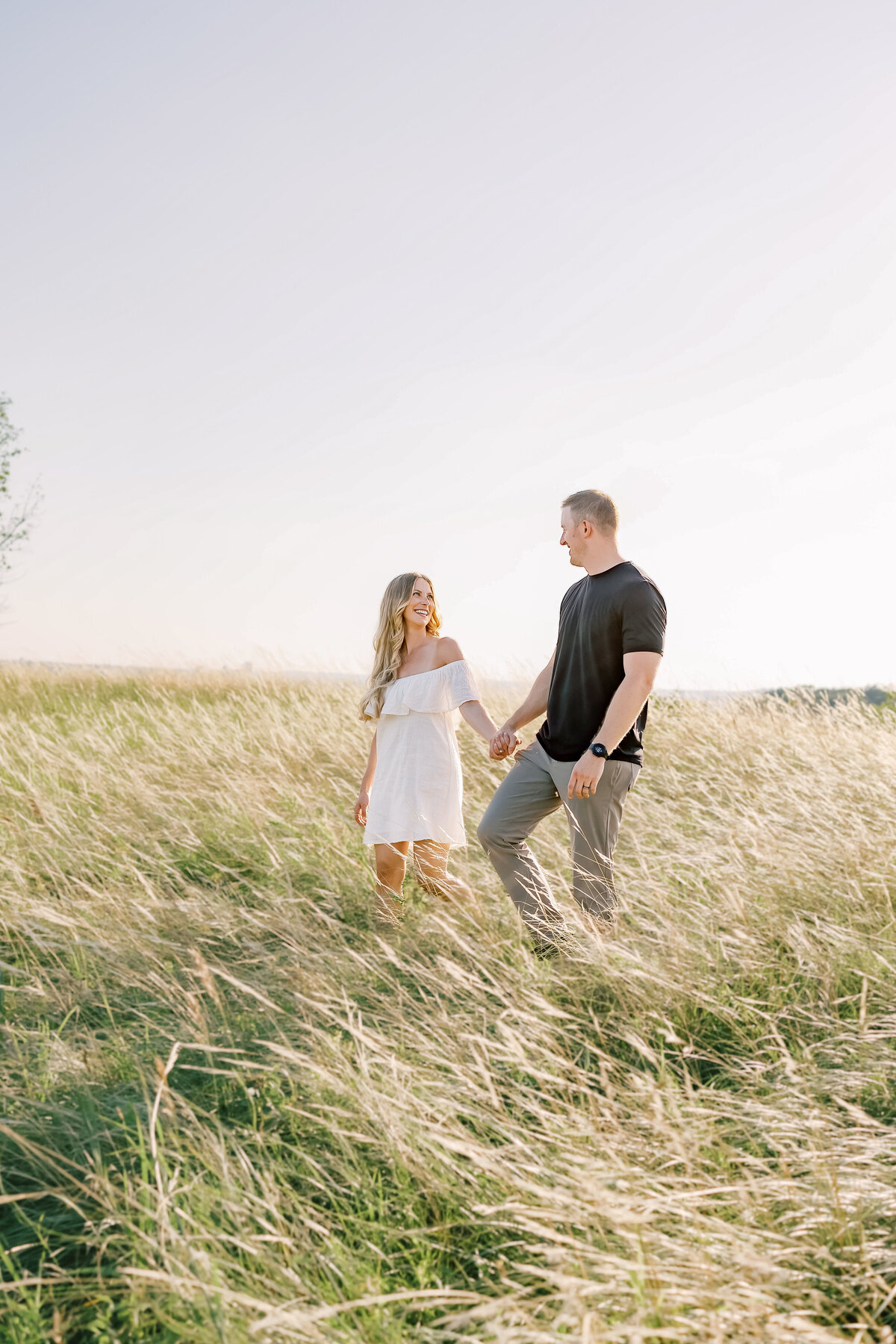 a joyful couple holds hands as they walk through a golden grassy field with soft sunlight. The couple smiles at eachother as they walk.