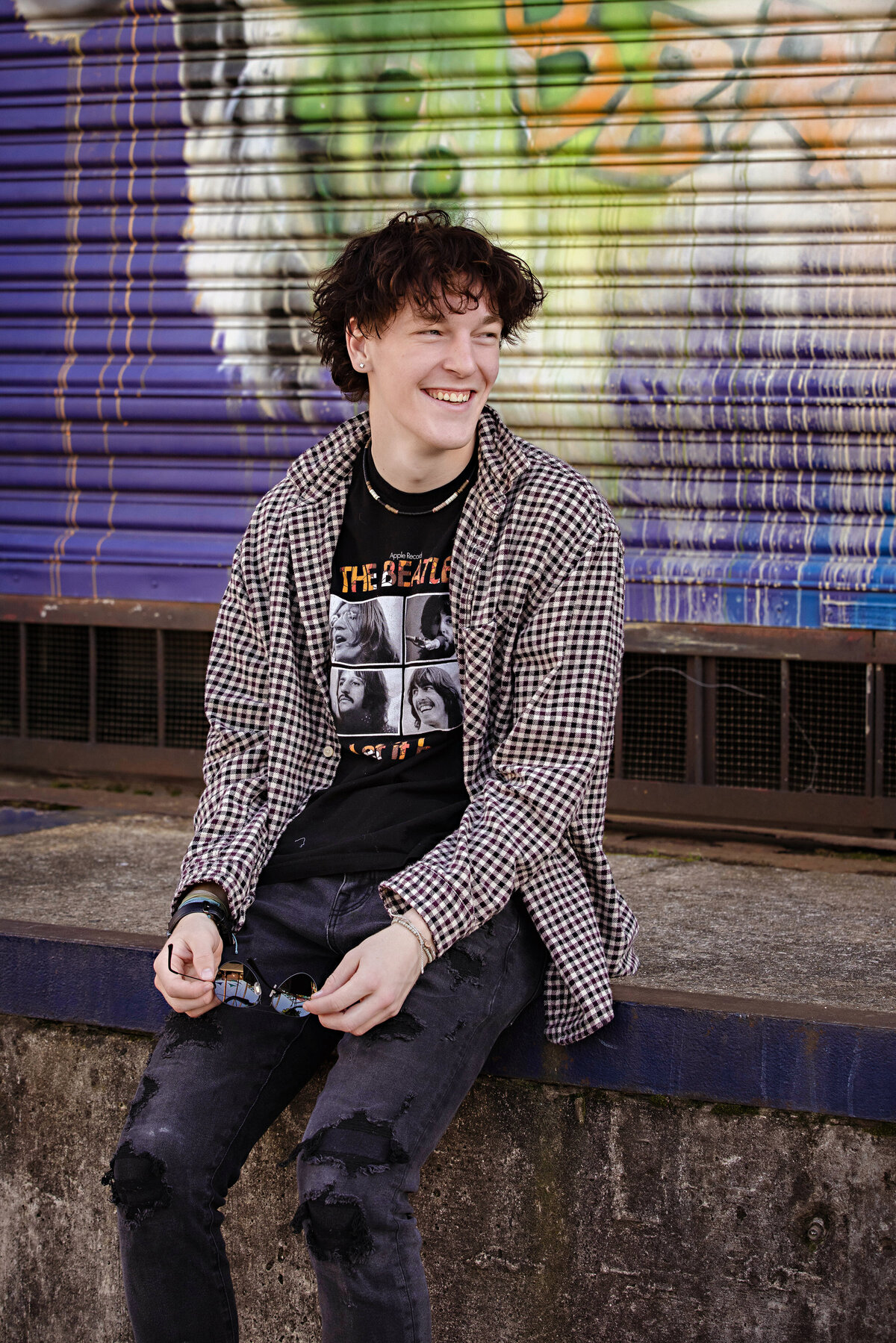 High school senior wearing a black Beatles t-shirt and checked button down, posing at Camp North End’s mural-lined loading dock in Charlotte NC, captured by Charlotte senior photographer.
