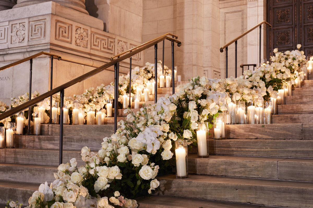 Here is the stunning photo taken at New York City, precisely at The New York Public Library by the talented photographer Christian Oth with East West Films as Videographers. The image captures a grand staircase adorned with white roses and other white flowers, gracefully cascading along the steps. To complement the floral display, lit candles are artfully placed among the blooms, imbuing the scene with a romantic and elegant ambiance. Additionally, the background showcases intricate architectural details, adding a touch of architectural splendor to the overall composition.