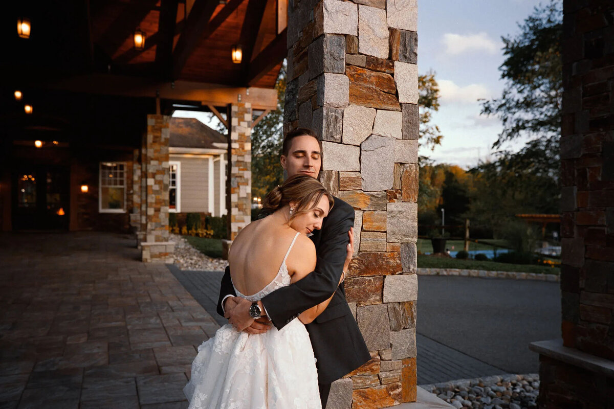A bride and groom embrace tenderly by a stone pillar at sunset, captured beautifully by an NJ wedding photographer. The bride in white rests her head on the groom’s chest as he gently hugs her, eyes closed.