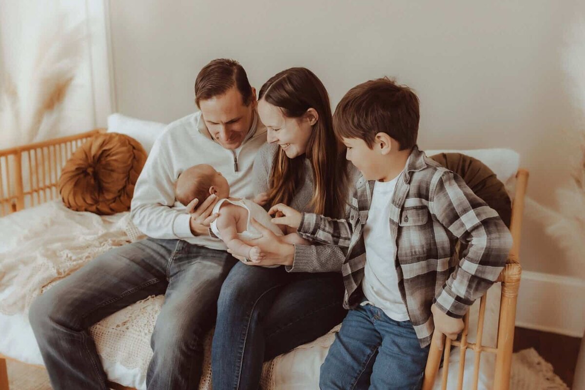 A family of four sits on a bed; the parents smile at a baby as an older child in a plaid shirt looks on, gently touching the infant—captured by a Macon studio newborn photographer.