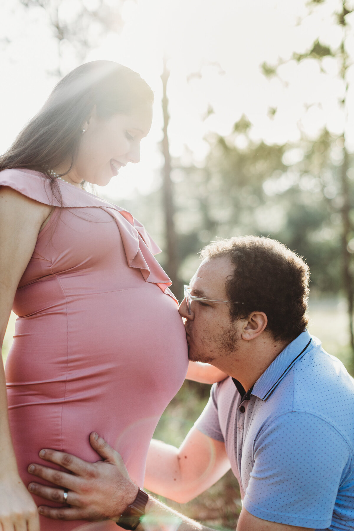 Expecting mother in a pink dress smiling as her partner kisses her baby bump during a soft, natural-light maternity session in Winter Garden, Florida