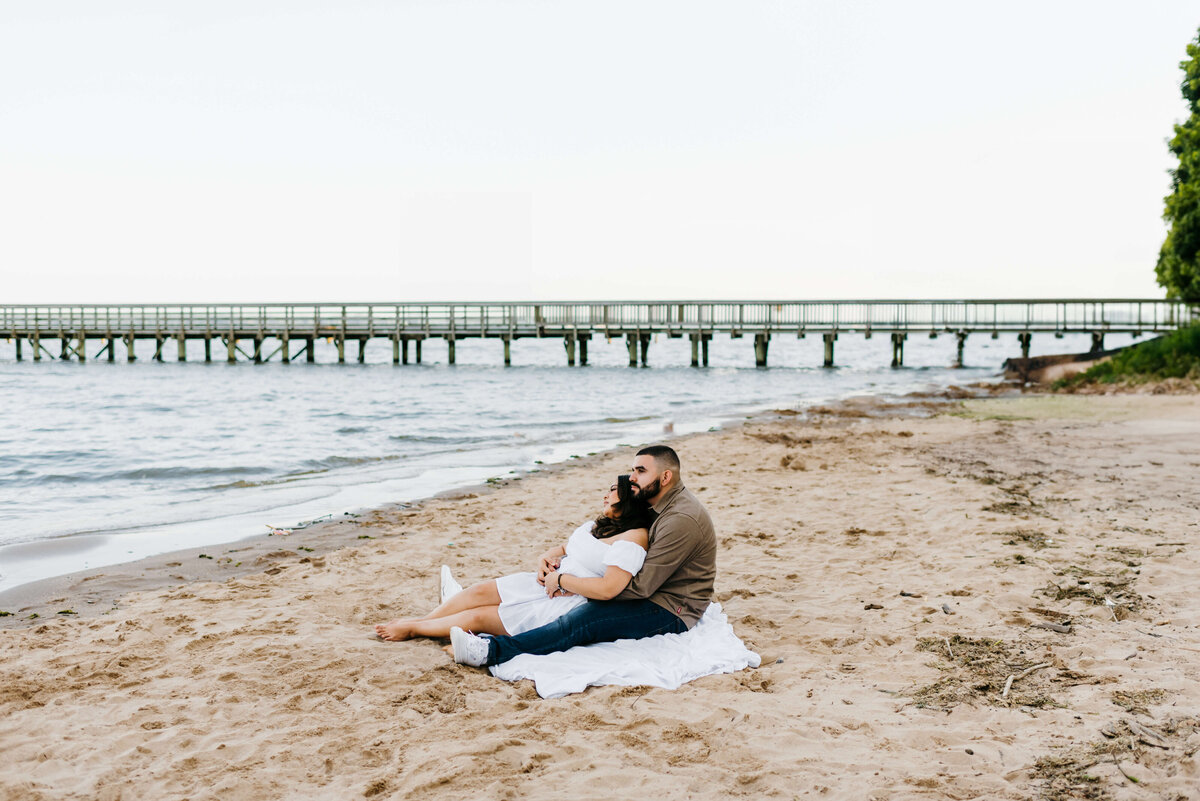 couples photography by Virginia wedding photographer with man and woman sitting together on a white blanket on the beach looking out at the water with a long dock over the water behind them