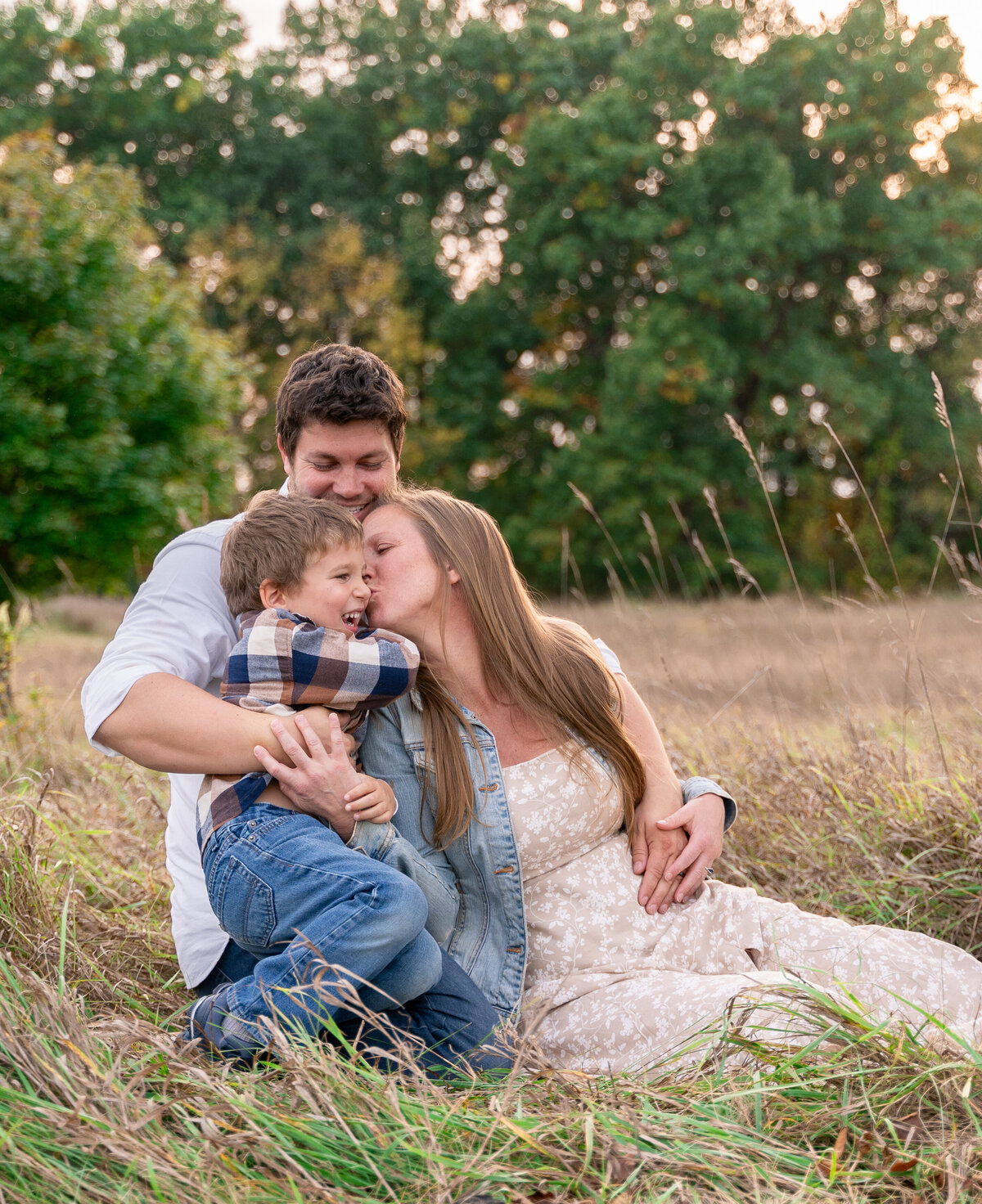 dad hugging mom and son while they all are sitting on the ground in an open field with the sunset in background 