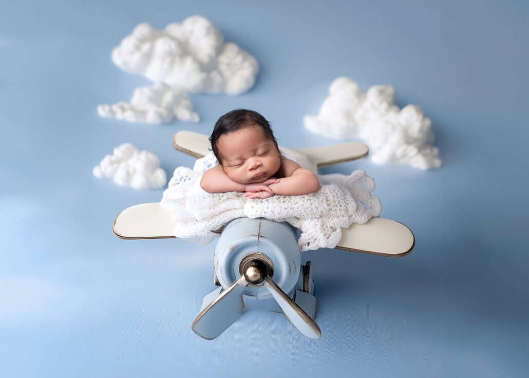 Newborn photography -- baby sleeping on a small blue airplane prop with white crochet blanket and fluffy cloud backdrop in a Birmingham photography studio