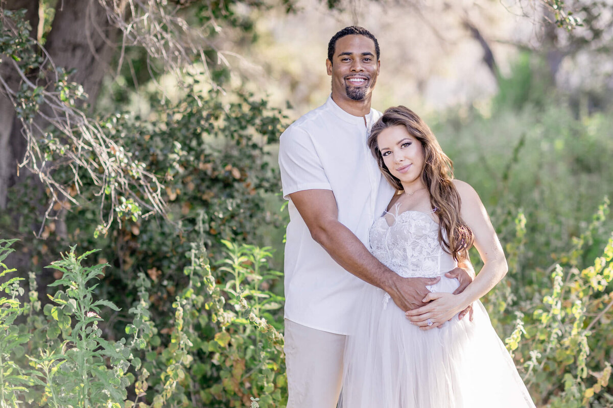 A couple stands in a sunlit, lush green outdoor setting. The man in a light shirt gently holds the woman, who is wearing a white dress. Both are smiling softly amidst the natural backdrop of trees and foliage.