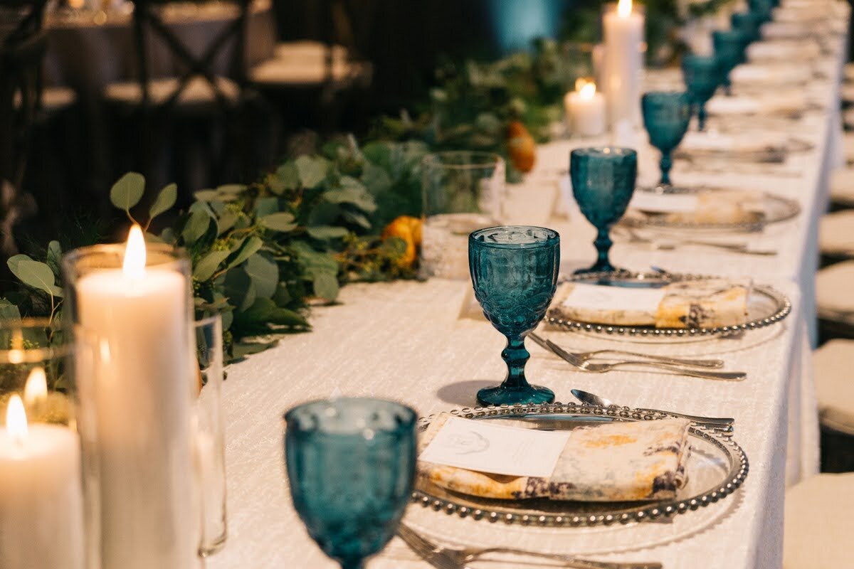 Wedding reception with candles, greens, place settings, and blue glass goblets.