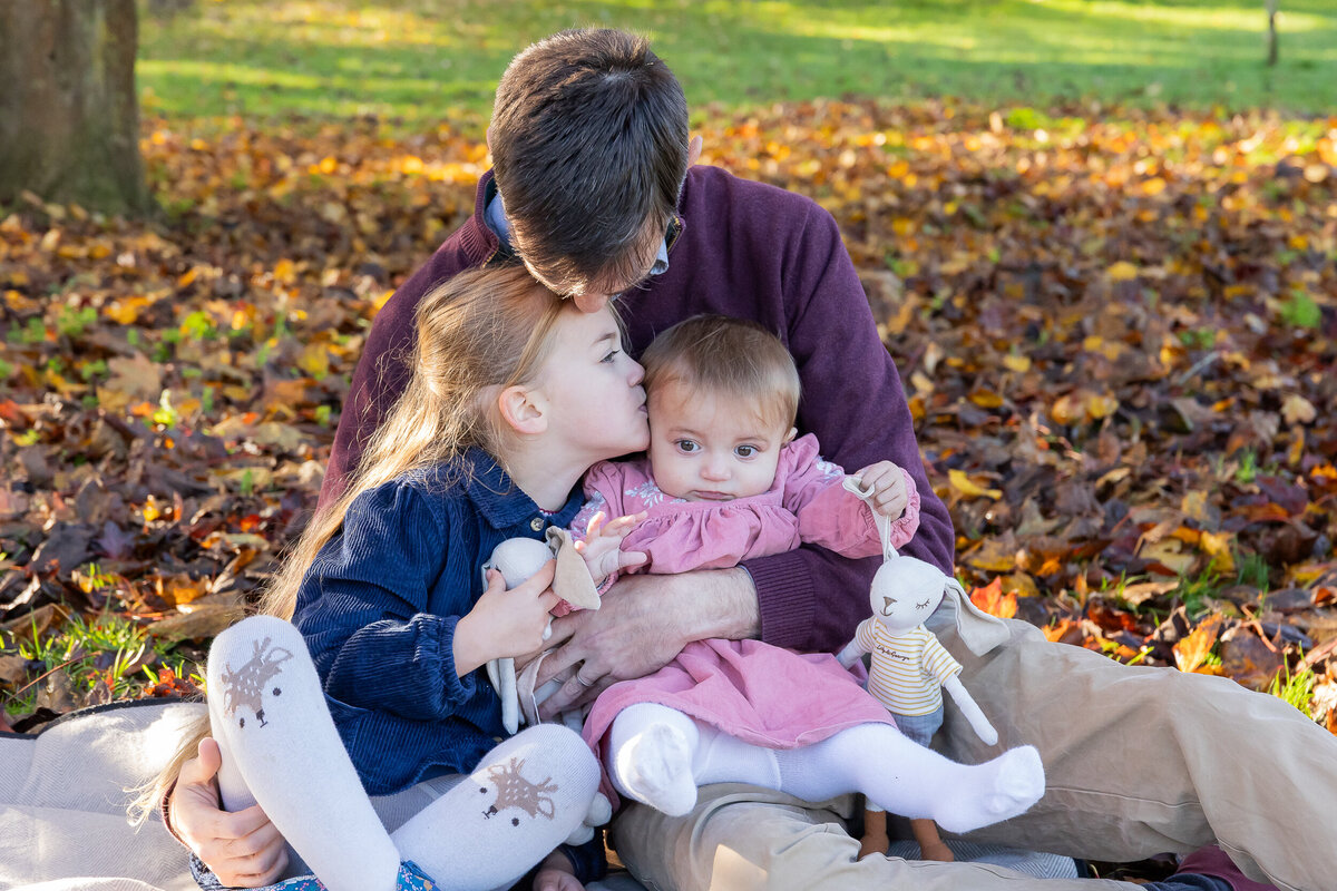 A father kisses his toddler daughter who gently kisses her baby sibling in a park with autumn leaves around.