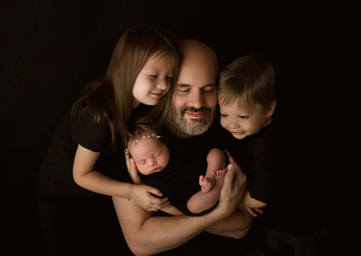 Dad and kids hugging newborn on black backdrop - heartwarming sibling portrait in Minneapolis studio 