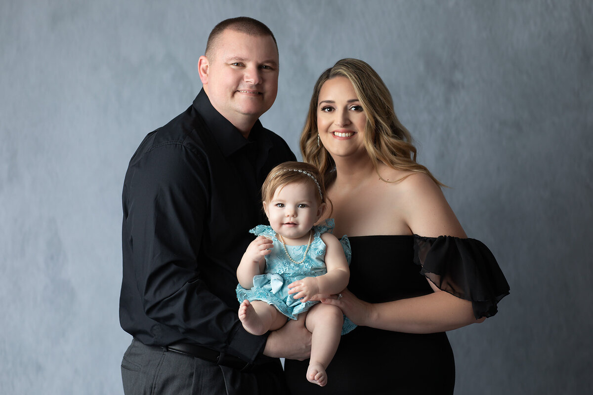 A smiling family of three poses in the studio, with their baby in a blue lace romper sitting between them, captured by a DFW milestone photographer.
