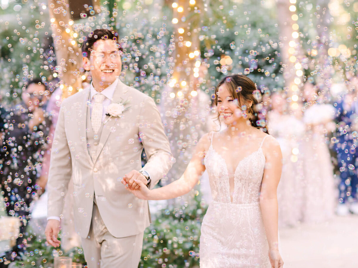 A joyful couple in wedding attire walks hand in hand through a flurry of bubbles in a garden setting.