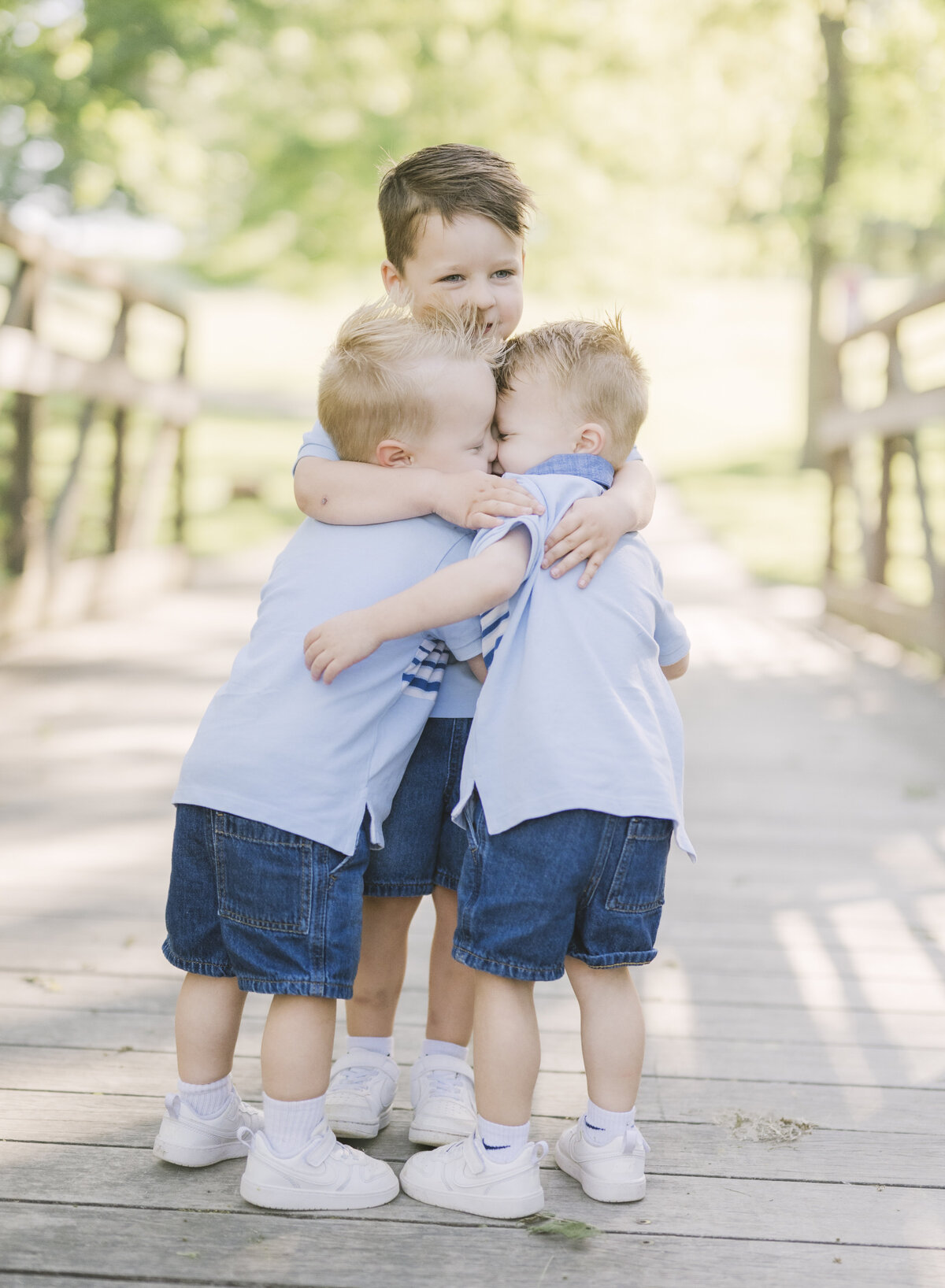 Three brothers hugging tightly on a wooden bridge in coordinated blue outfits during a Kansas City milestone photo session.