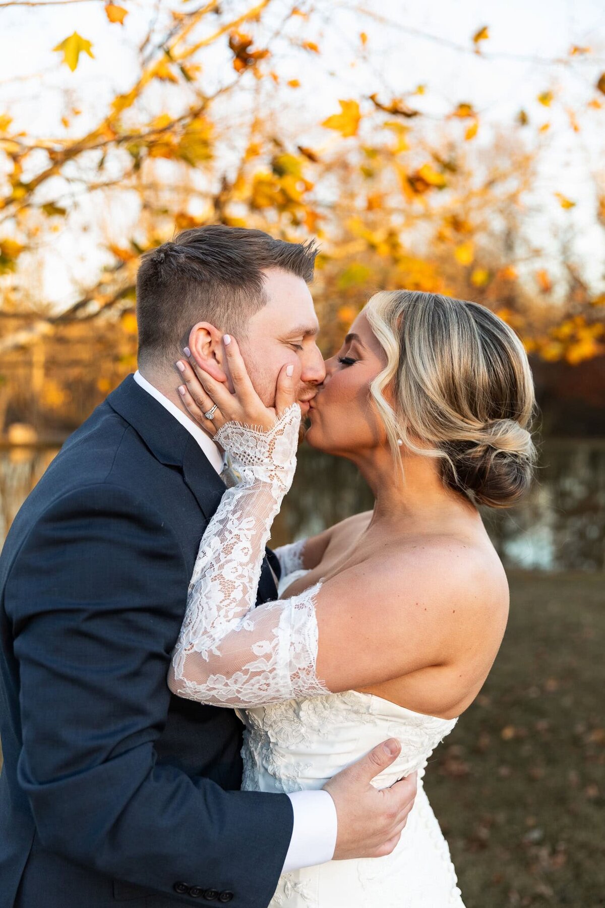 The bride and groom share a tender kiss beneath a canopy of amber leaves, their hands pressed together in quiet devotion. Her off-the-shoulder wedding dress, adorned with delicate lace sleeves, complements the groom’s dark suit, creating a perfect contrast against the warm autumn tones of their Pennsylvania wedding day