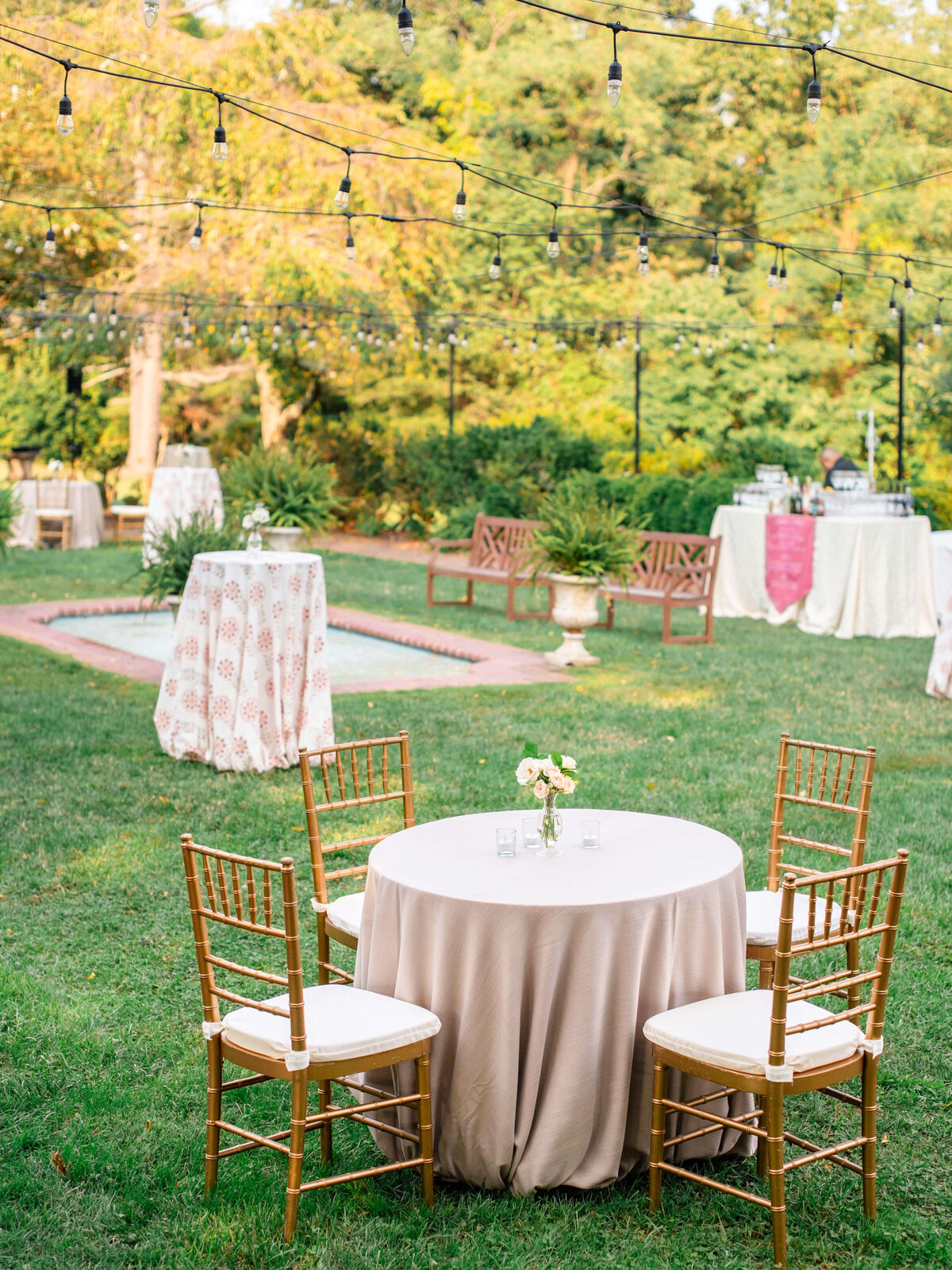 A garden setting features a round table with a beige tablecloth and four wooden chairs. String lights hang above, and there are additional tables and lush green trees in the background, creating a serene outdoor event atmosphere.