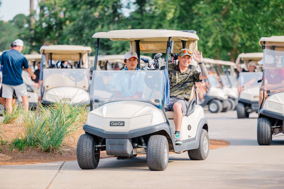 A group of people in multiple golf carts cruises along a paved path, surrounded by greenery. Captured by an event photographer in Fayetteville, NC, a person in the front cart waves cheerfully while the passenger sits beside them. Other carts and people can be seen in the distance.