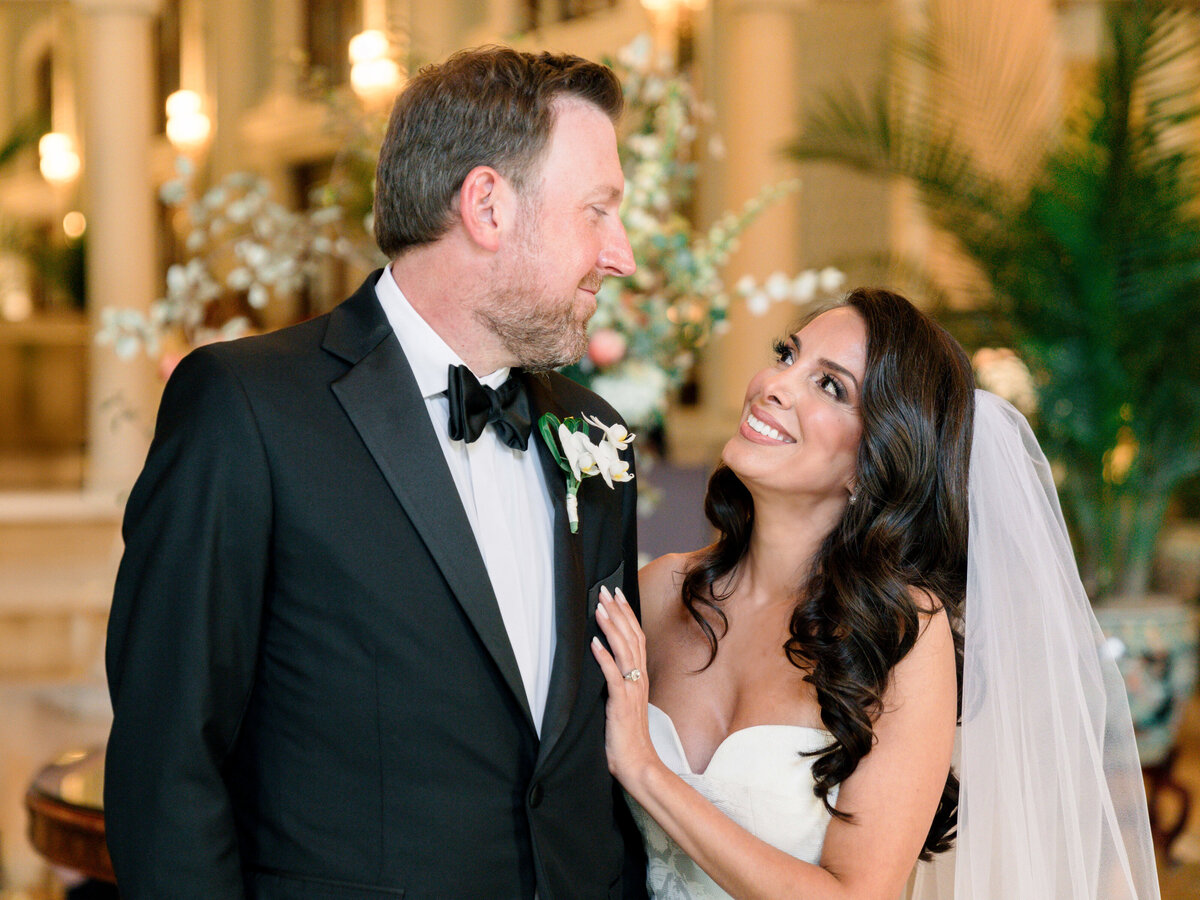 A bride and groom share a kiss in a luxurious, ornately decorated hallway. She wears a long white gown and veil, holding a bouquet. He is in a black tuxedo. The setting features elegant chandeliers and intricate ceiling designs.