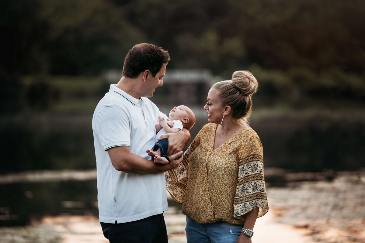 new parents holding and looking lovingly at their infant  at sunset in the summer in front of a lake