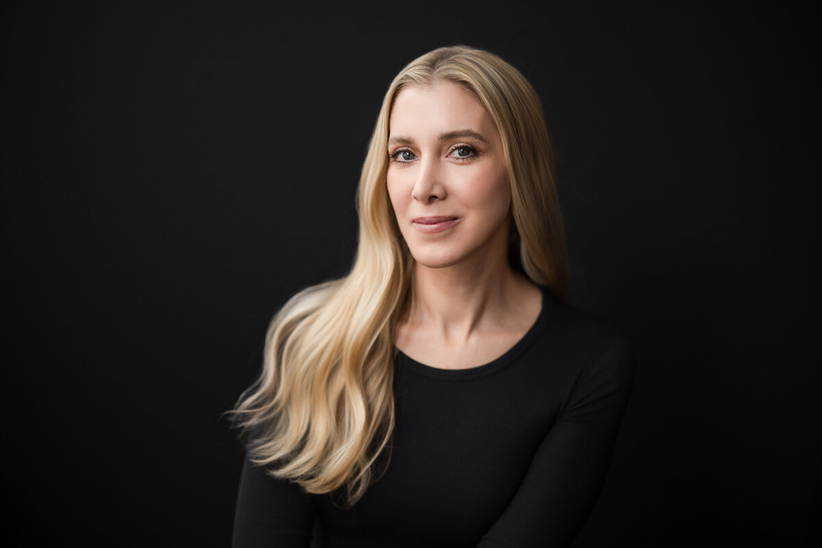 Person with long, blonde hair wearing a fitted black top, seated against a dark background. They are softly smiling and looking into the camera with a calm, confident expression. The lighting is clean and natural, highlighting their features in a modern, minimal portrait style.
