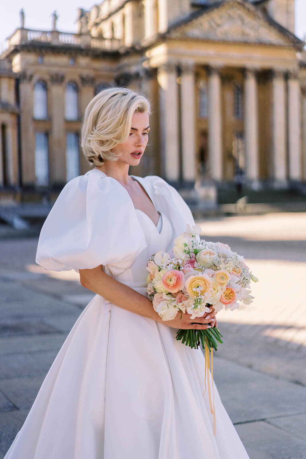 luxury bride standing in front of blenheim palace wearing a dress with large puffy sleeves and holding a peach wedding bouquet