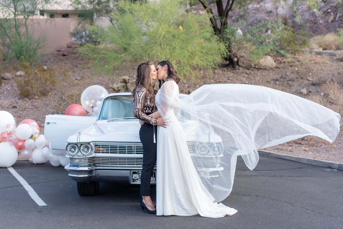 Two brides kiss in front of a vintage car with a veil blowing in the wind