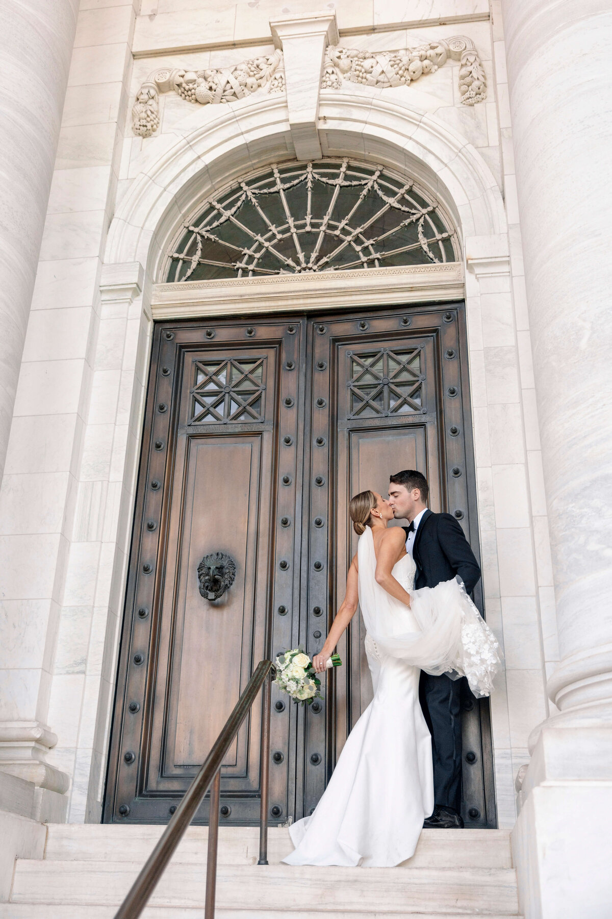 A bride and groom share a kiss in front of large, ornate wooden doors. The bride holds a bouquet of white flowers and is in a white gown, while the groom is in a black suit. They are on a marble staircase, surrounded by elegant architectural details.