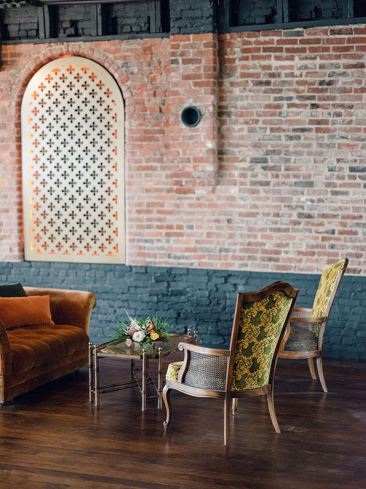 The Little Bird Lounge at Clementine Hall with vintage soft seating. A velvet rust colored sofa and twin set of gold and blue arm chairs around a mid century brass and glass coffee table with a small tropical flower centerpiece.