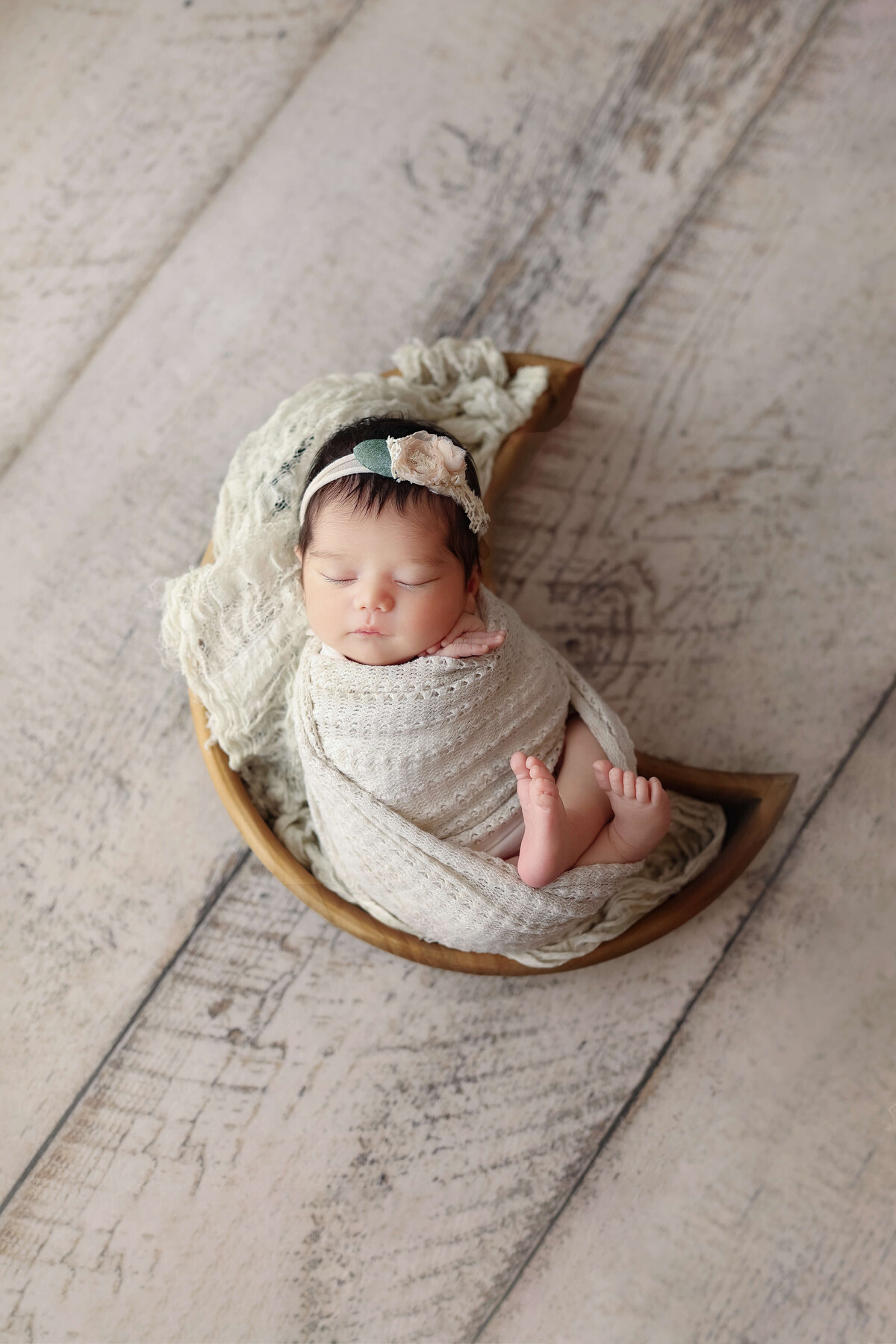 baby girl in a white swaddle laying in a moon shaped wooden bowl on a white wood floor