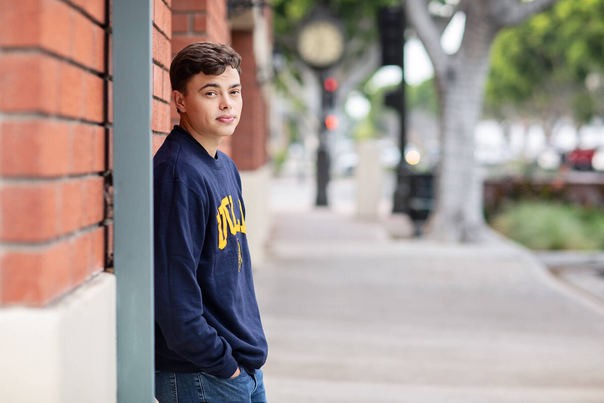 A young man with brown hair leaning his back against a brick wall, wearing a navy crewneck and looking over his right shoulder at the camera, barely smiling. Captured in Old Town Tustin.