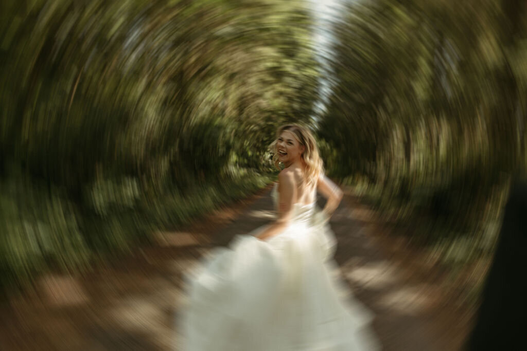 A bride joyfully frolics through a lush green forest along a dirt path in her elegant wedding dress.