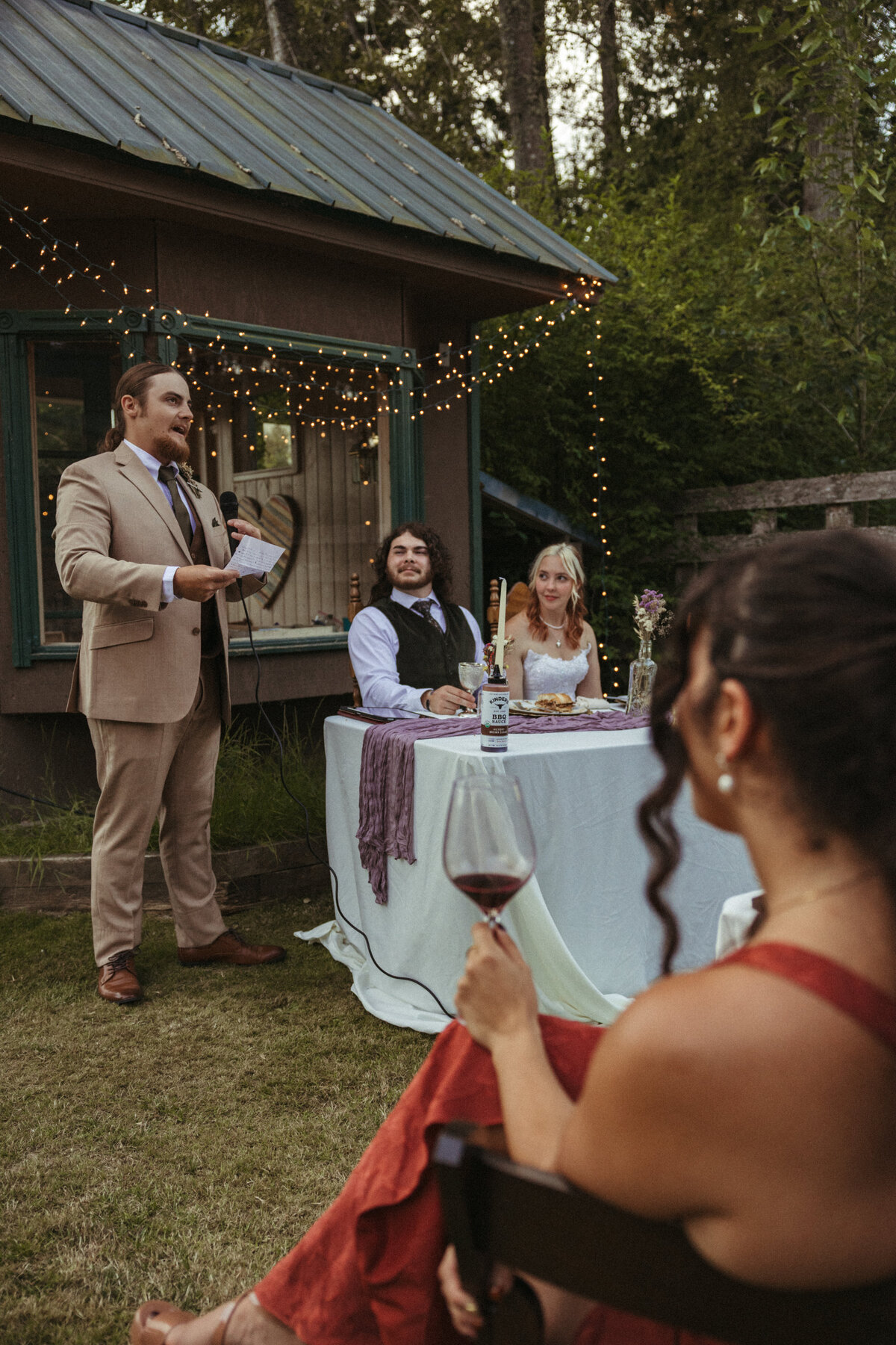 The best man gives a speech during an intimate backyard wedding, beneath a forest donned with twinkle lights and towering trees overhead.