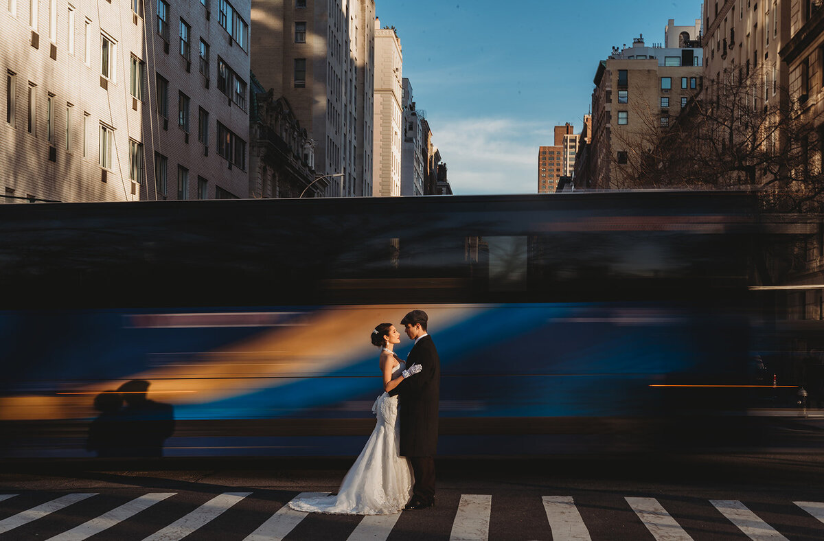 bride and groom stand together as bus blurs by in background on NYC street crosswalk photo by cait fletcher photography