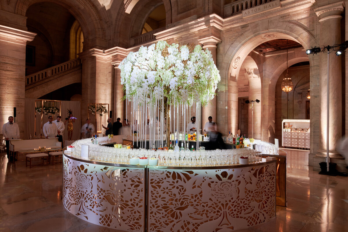 The captivating photo was taken at the iconic New York City location, The New York Public Library, by the talented photographer Christian Oth. It showcases a stunning event setup with a circular bar embellished with intricate floral patterns at its base. Above the bar, exquisite white flower arrangements cascade gracefully, adding a touch of elegance to the scene. The grand hall, with its arched architecture and soft lighting, provides a luxurious backdrop for attendees and staff to gather and immerse themselves in the opulent ambiance.  Videographers: East West Films