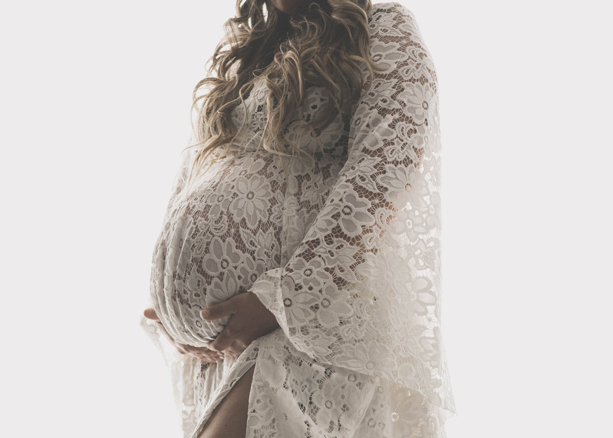 A mother to be holds under her bump while standing in a lace gown