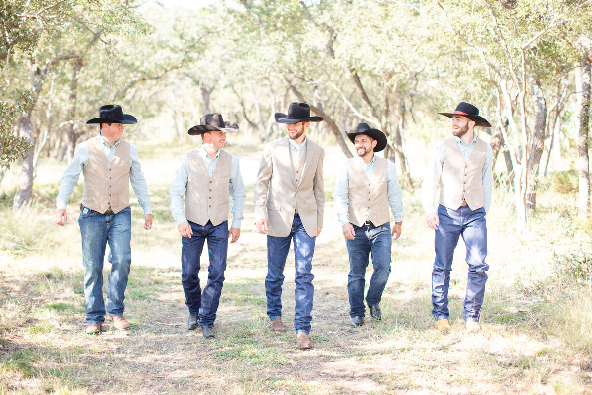 groomsmen in tan vests jeans and black cowboy hats walk to the Hill Country wedding venue by San Antonio wedding photographer Firefly Photography