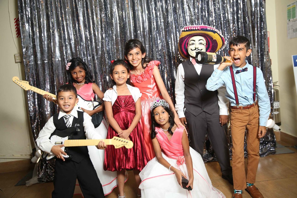 Young children pose in front of silver backdrop wearing masks and hats and holding fun props. Photobooth by Ross Photography, Trinidad, W.I..