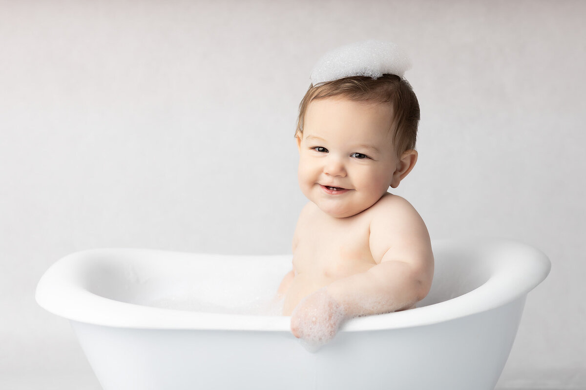 Dallas milestone photographer captures a baby with bubbles on their head, smiling in a white vintage tub during a light and airy studio bath session.