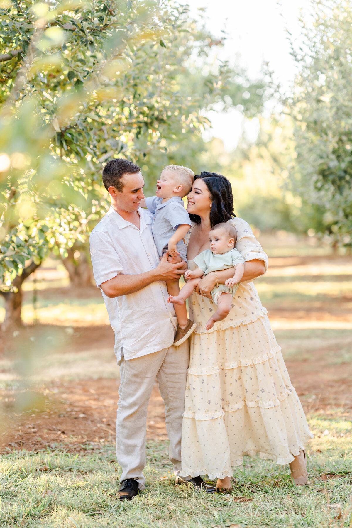 A mother dressed in a yellow floral gown playfully snuggles her toddler son as she holds her baby and her husband smiles lovingly at his family. The family stands in a orchard field and was photographed by Bay Area Family Photographer, Light Livin Photography.