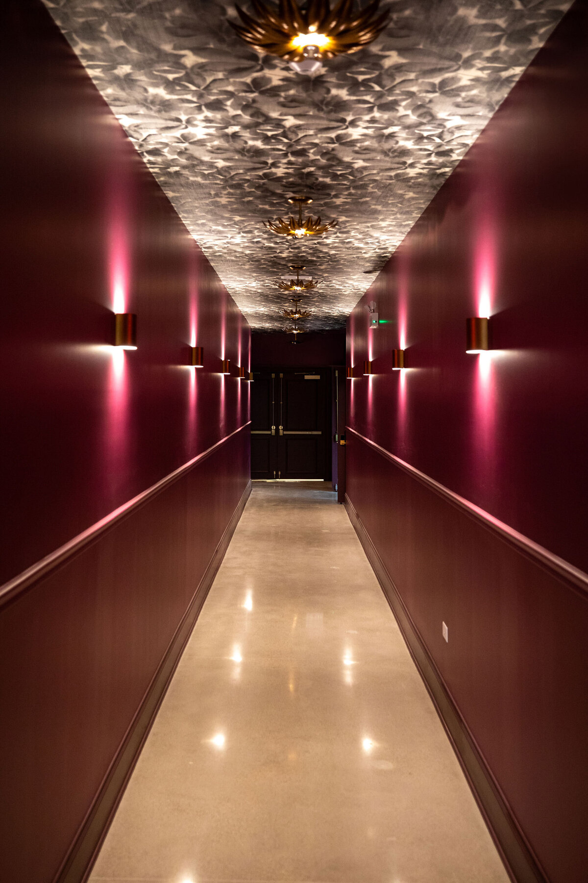 Striking venue hallway with dramatic lighting, vibrant wall color and ceiling wallpaper, sleek flooring, and blend of modern and antique lighting accents creating a bold, stylish atmosphere