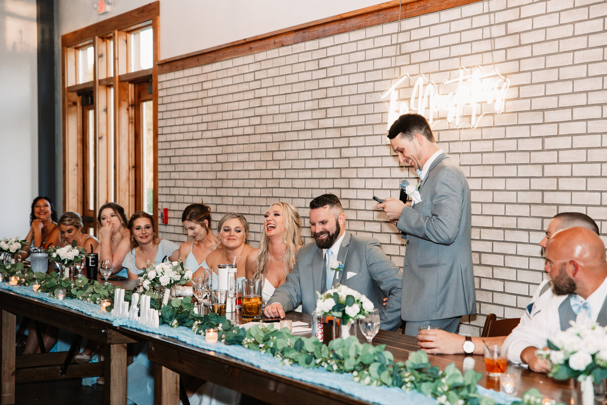 A groomsman gives a funny toast at the long head table to the laughing bride and groom
