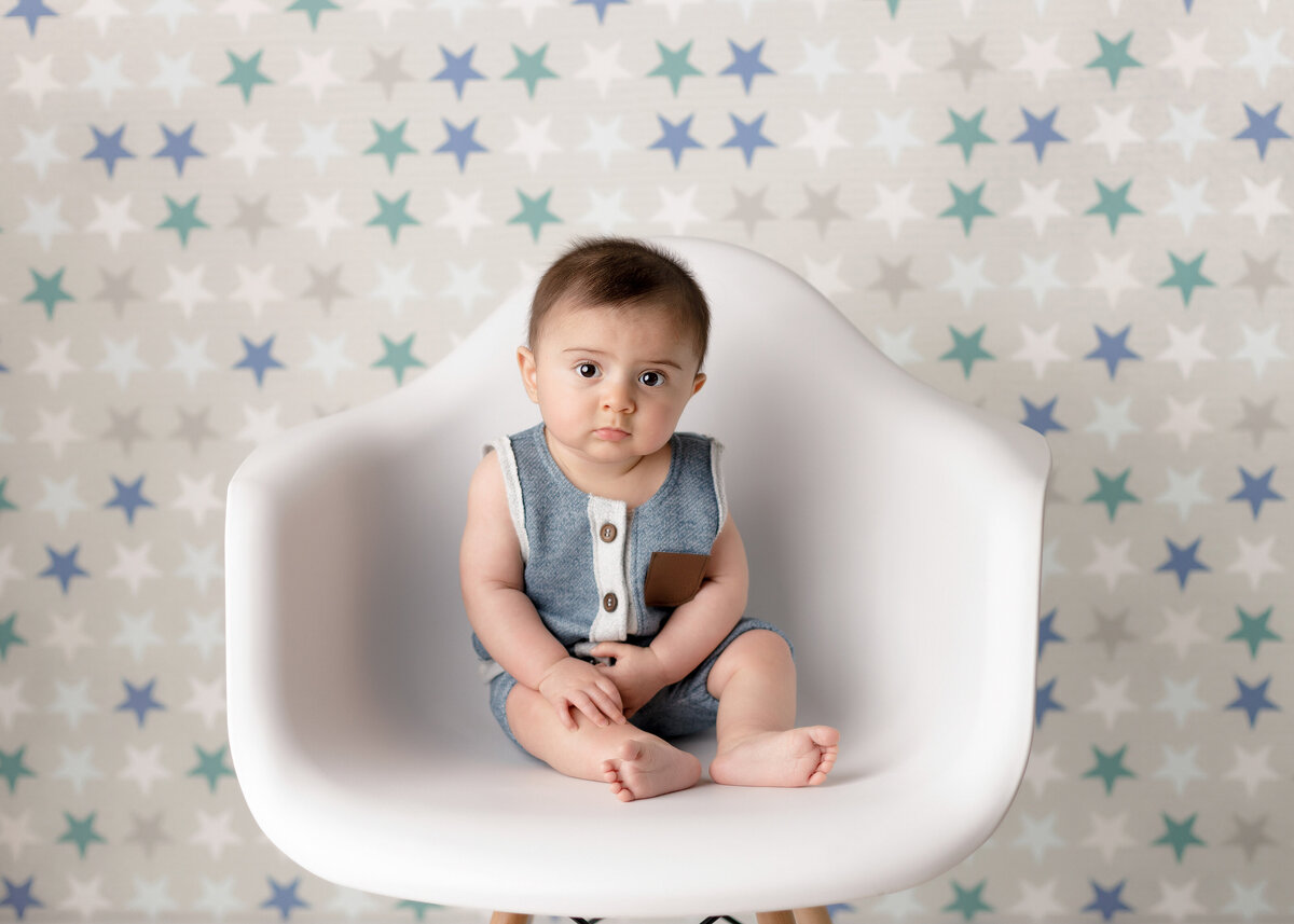 6-month baby photoshoot at West Palm Beach and Delray Beach baby photographer.  Baby is sitting on a white Eames chair in a blue and white jumper. Baby is looking at the camera with a serious expression. The backdrop is of white, blue, and green stars.