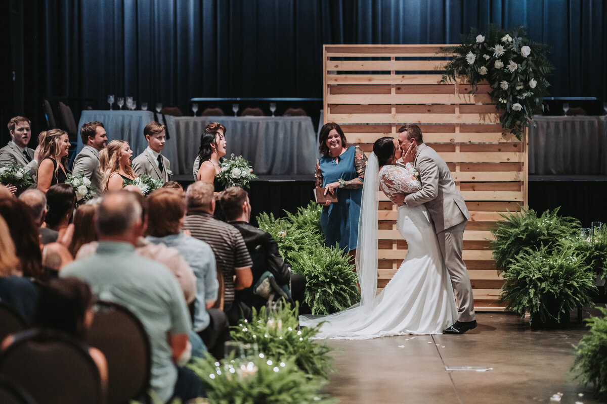 Indoor wedding ceremony with wooden backdrop and floral decorations as guests look on in awe.
