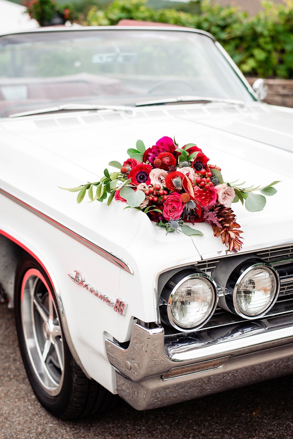White vintage 1964 Oldsmobile with dark red leather interior with large bridal bouquet featuring a cascade of amaranthus, red roses, blush roses, red anthurium, red anemones, protea, blue thistle, hypericum berries, dianthus and assorted greenery wrapped in an ivory satin ribbon at Arrington Vineyards.