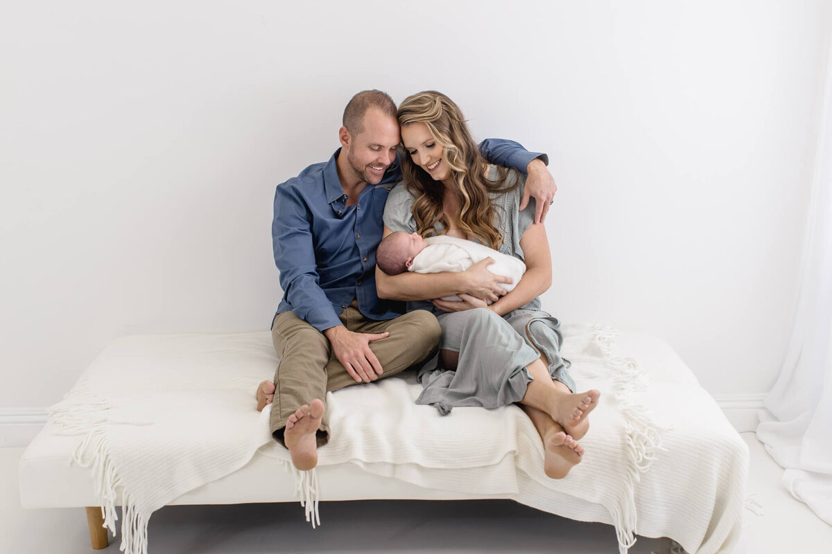 A couple sits closely on a white couch, smiling while holding a sleeping baby wrapped in a white blanket. They are dressed in casual clothing, and the setting is softly lit with a serene, minimalist background.