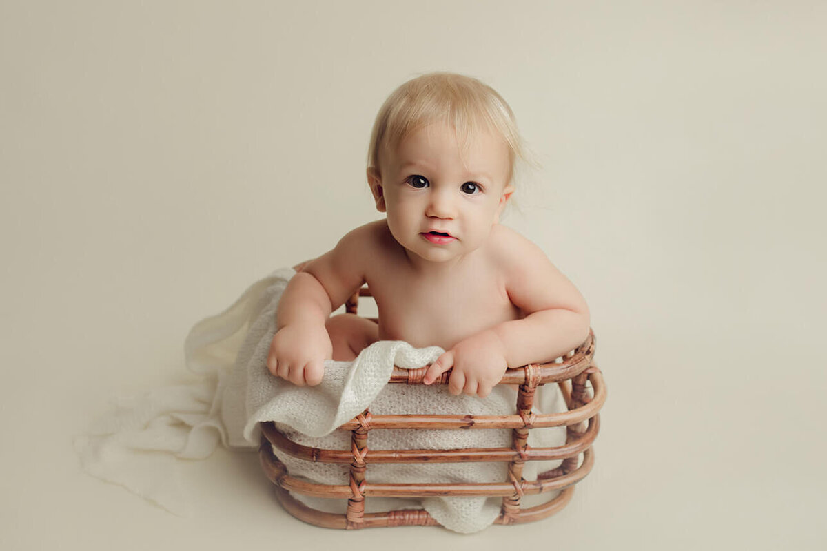 a nude one year old boy sitting in a basket on a white drop