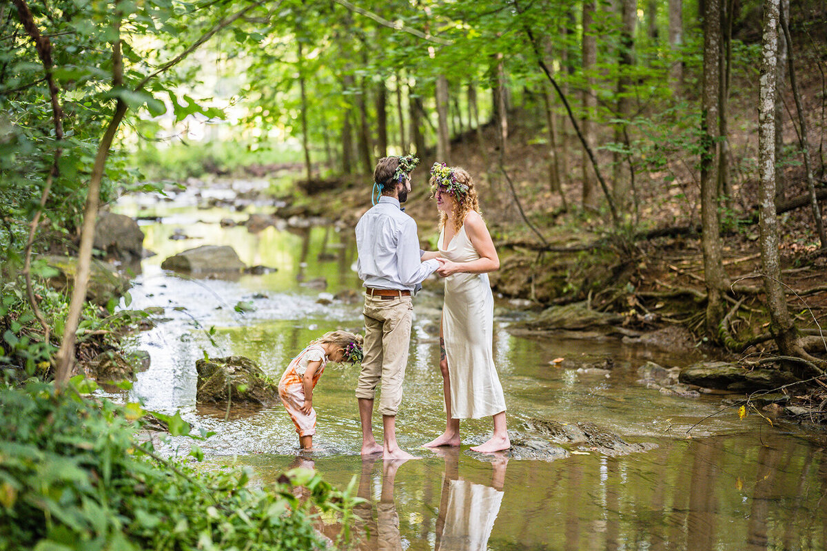 A bride and groom on their elopement day read their vows while standing in the creek at Fishburn Park. Their child can be seen behind the groom observing her reflection in the water.