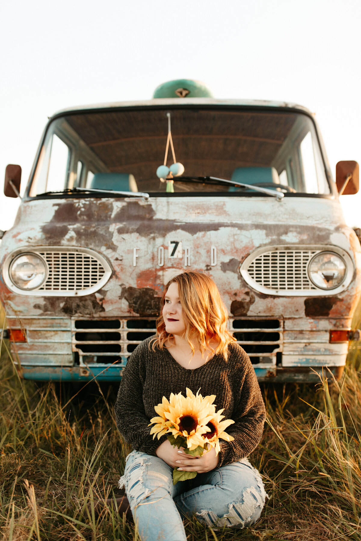 High school senior sitting on the ground in front of an old vintage hippie van and holding sunflowers