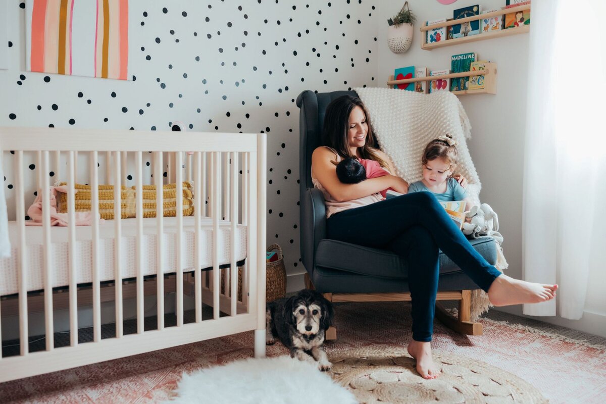 newborn mother and toddler reading while holding newborn in rocking chair in nursery with dog nearby by Orange County photographer Francesca Marchese