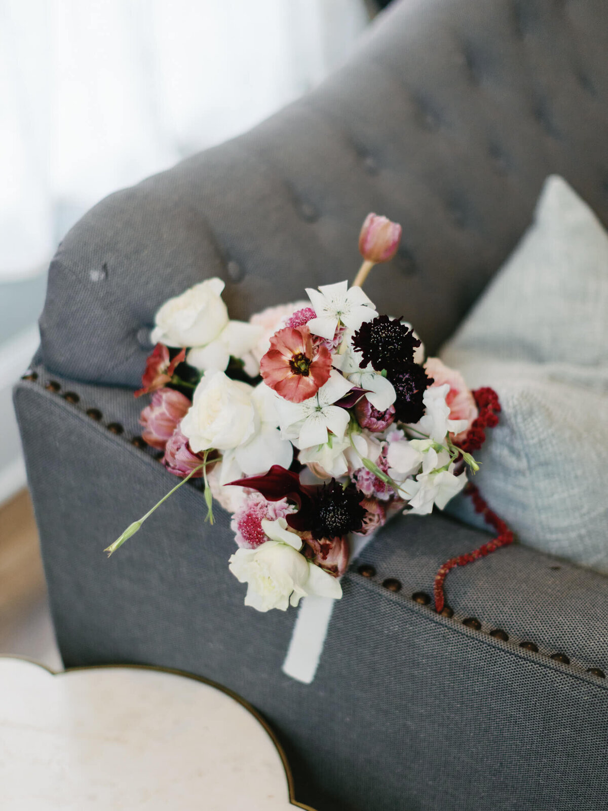 A vibrant bouquet of pink, white, and dark flowers rests on a grey tufted sofa.